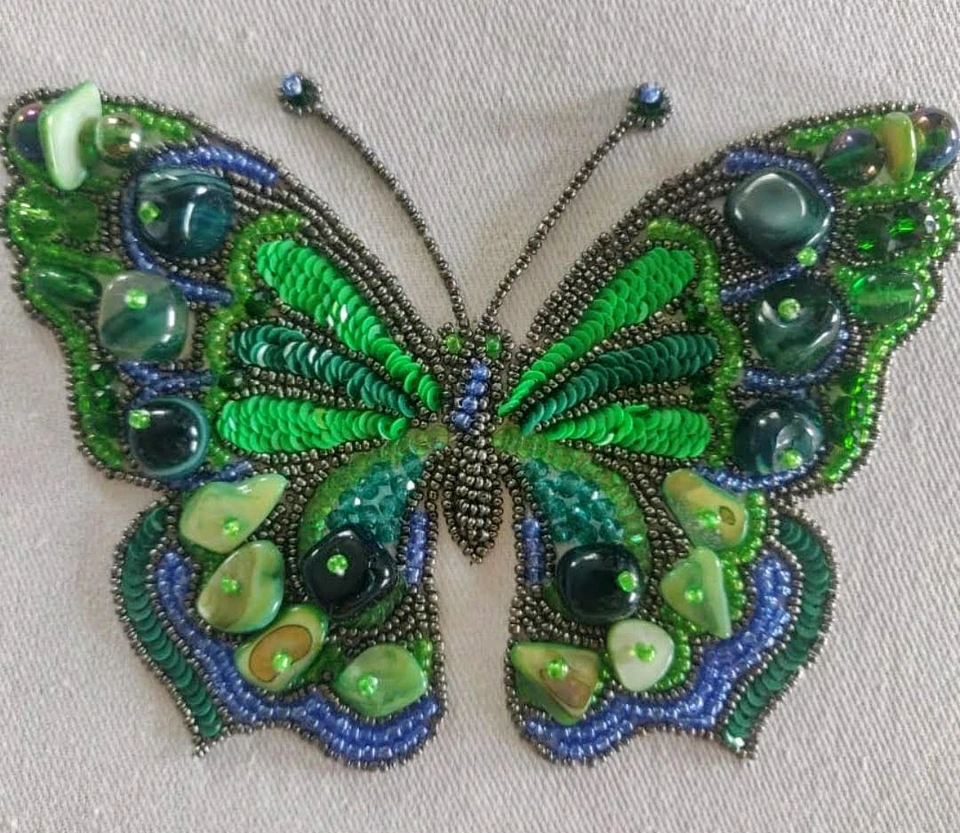 A green and blue butterfly embroidered with beads, sequins and reflective accents on pale fabric.
