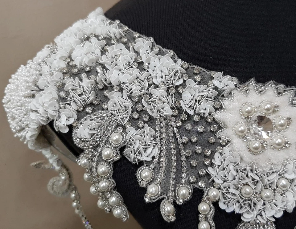 Close view of white floral embroidery with pearl-like beads, silver-toned details and hanging bead strands on a shaped garment panel.