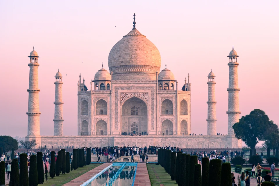 The Taj Mahal, its reflecting pool and visitors beneath a pink sky.